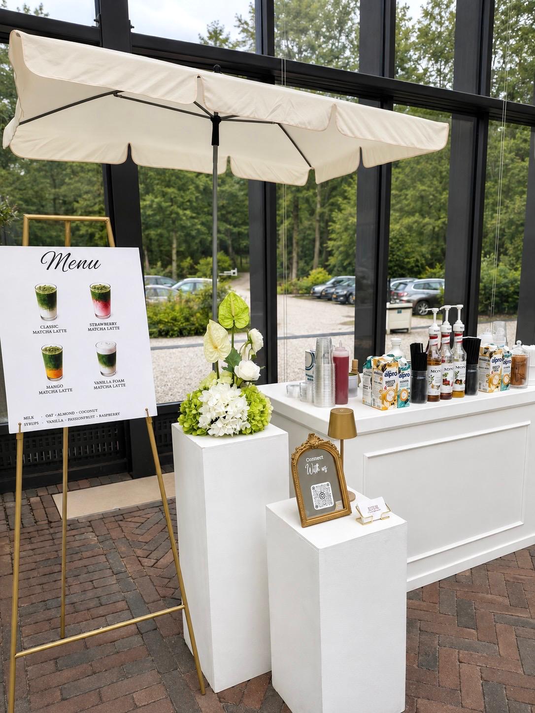 Matcha bar met parasol, menubord, siropen en bloemstuk op witte sokkels