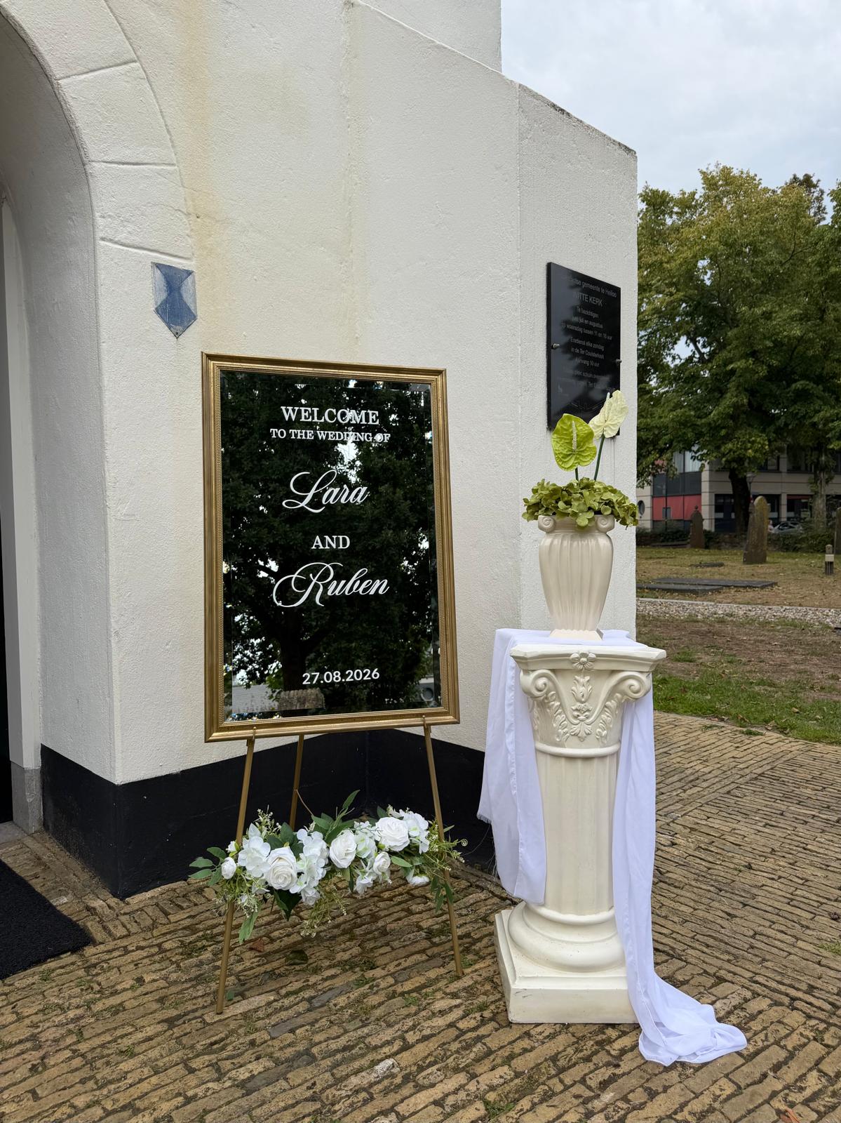 Spiegel-welkomstbord in gouden lijst bij de kerk met witte bloemen en zuil
